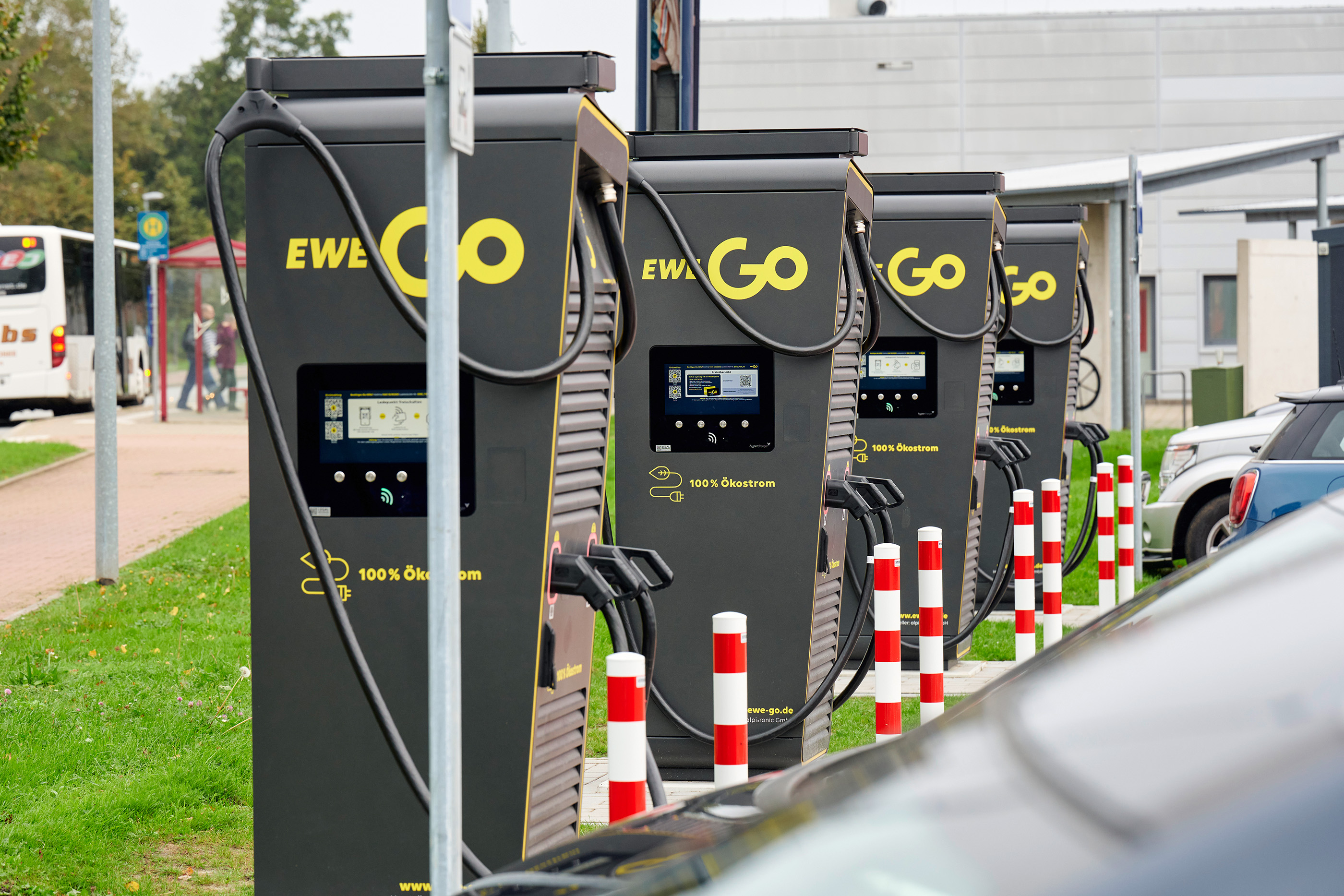 Das Bild zeigt eine Reihe von Ladestationen für Elektrofahrzeuge, die mit “EWE Go” gebrandet sind und ein grünes Steckersymbol anzeigen. Die Ladestationen sind anthrazit mit gelber Schrift und haben digitale Displays. Am rechten unteren Bildrand ist ein Elektroauto zu sehen, was darauf hinweist, dass sich die Ladestationen in einem für das Laden von Elektrofahrzeugen vorgesehenen Parkplatz befinden. Dies spiegelt die wachsende Infrastruktur zur Unterstützung von Elektrofahrzeugen wider, als Teil des Übergangs zu nachhaltigen Transportoptionen.