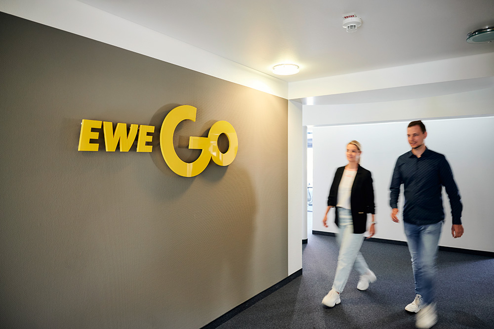 Zwei Personen laufen in einem Flur an einem EWE Go-Logo vorbei. Das Logo hängt an einer grauen Wand.