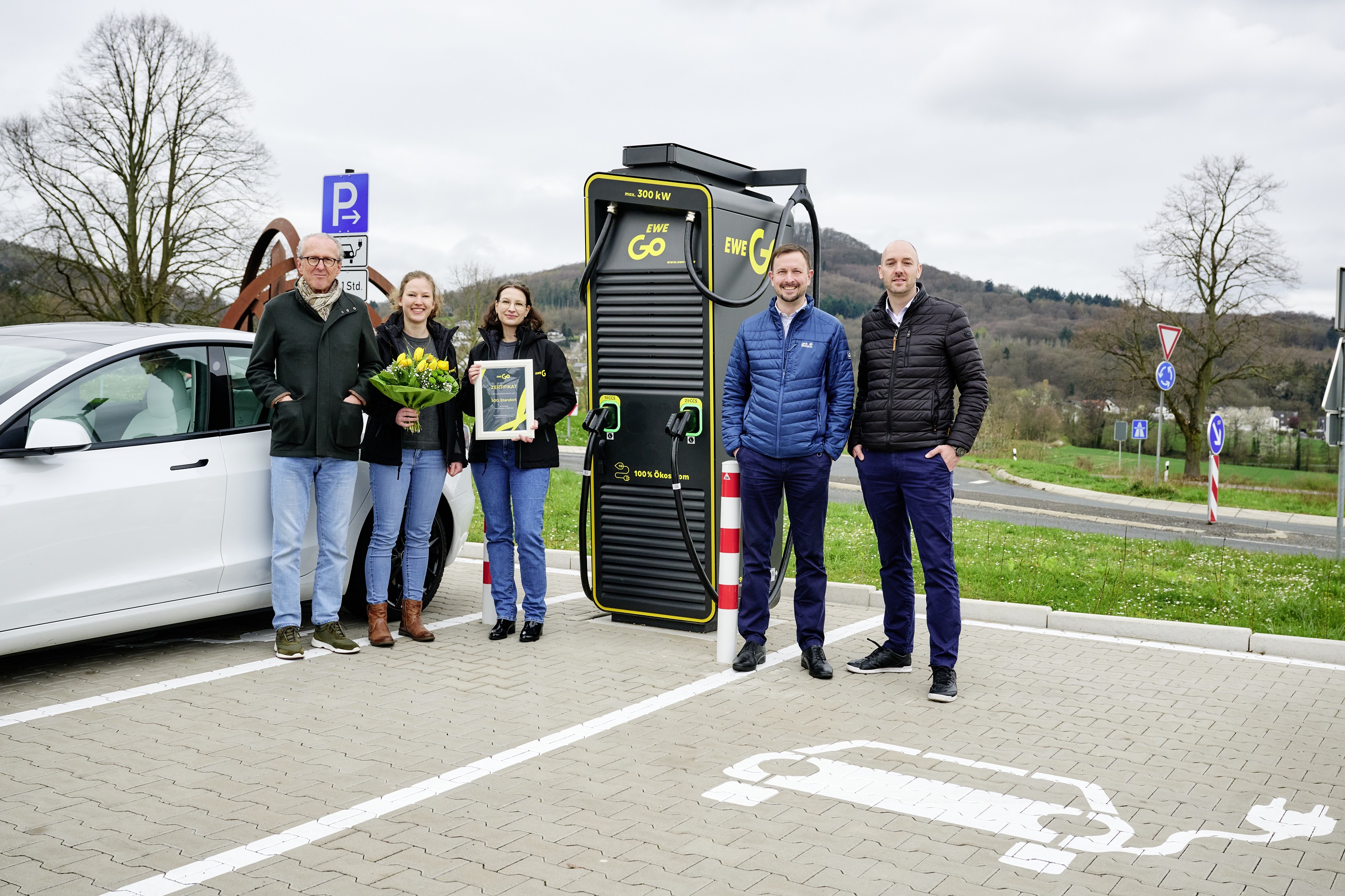 Im Zentrum des Bildes ist eine Schnellladestation von EWE Go zu sehen. Links daneben stehen 3 Personen, eine mit Blumenstrauß, eine weitere mit einem Bilderrahmen. Rechts neben der Ladestation stehen 2 Personen. Am linken Bildrand ist ein weißer Tesla zu sehen. Im Hintergrund befindet sich eine Wiese und Bäume.