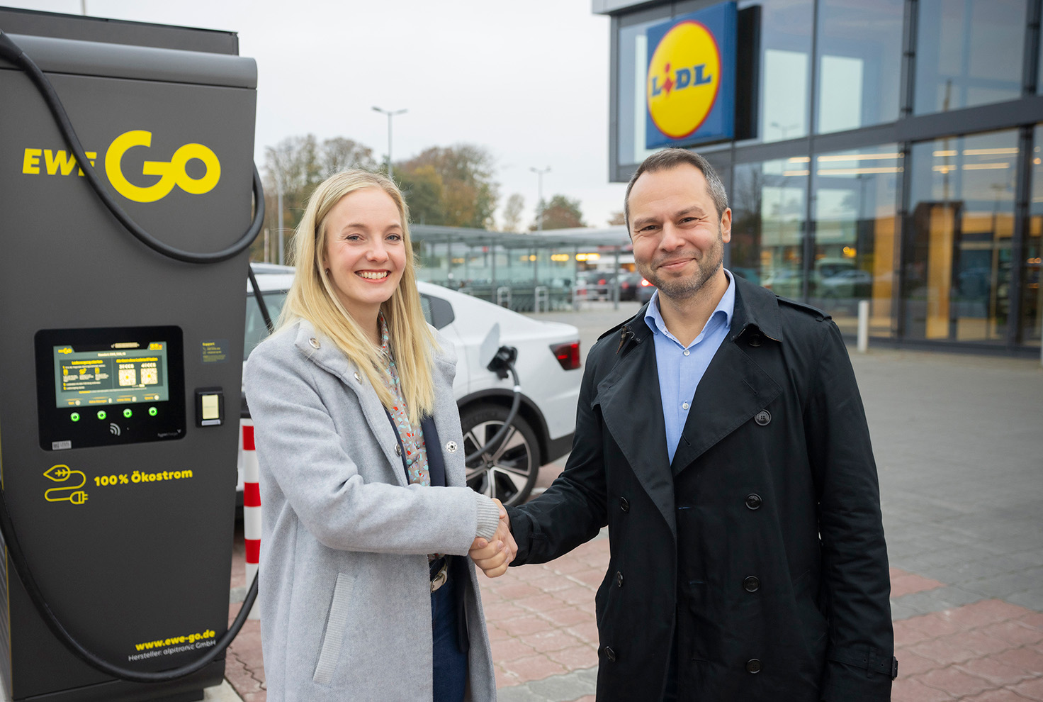 Jana Czeczor, Bereichsleiterin Immobilien, Lidl Immobilien Dienstleistung GmbH & Co. KG und EWE Go-Geschäftsführer Ilker Akkaya stehen vor einer EWE Go-Ladesäule auf dem Parkplatz einer Lidl-Filiale und schütteln sich die Hand. Im Hintergrund ist das Lidl-Logo gut sichtbar. Die Szene symbolisiert die Zusammenarbeit von EWE Go und Lidl für den Ausbau des Schnellladenetzes für E-Autos in Deutschland. Die Ladesäule zeigt das Logo von EWE Go und den Hinweis auf 100 % Ökostrom.