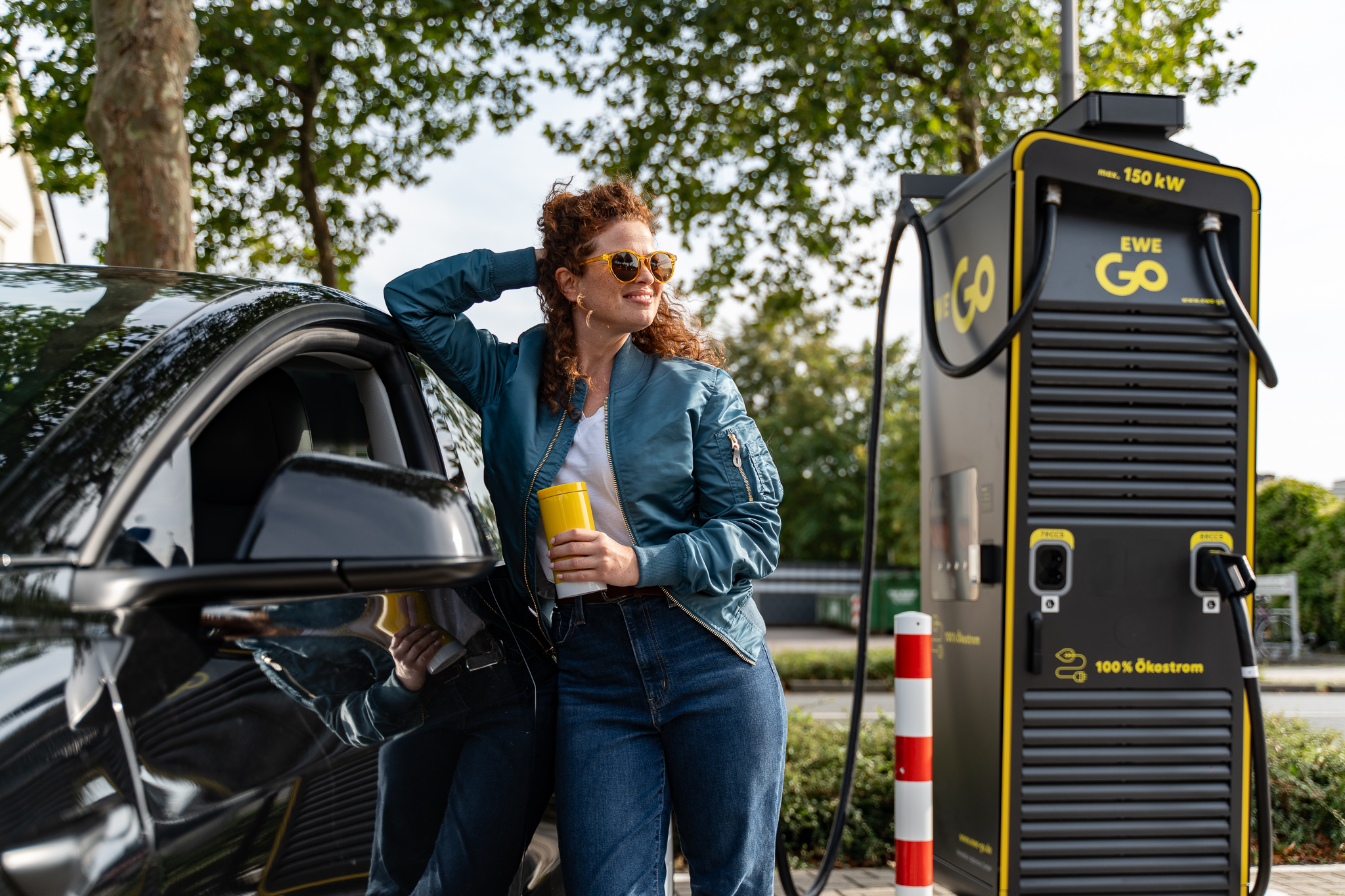 Das Bild zeigt eine Frau, die neben einem schwarzen E-Auto steht, das an einer Ladestation aufgeladen wird. Die Ladestation ist mit “EWE Go” und einer Leistung von “150 kWh” beschriftet. Zudem ist vermerkt, dass der Strom zu “100% Ökostrom” ist. Die Frau hält eine gelben Thermosbecher in der Hand. Dieses Bild unterstreicht die wachsende Infrastruktur für Elektrofahrzeuge und die Förderung umweltfreundlicher Energie.