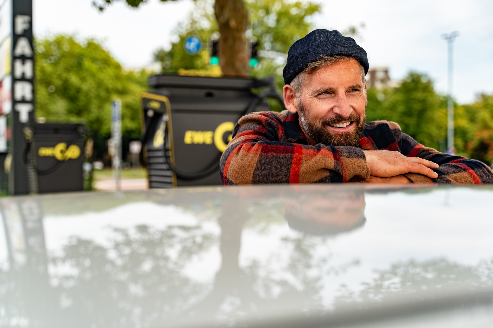 Ein Mann mit grauem Bart und einer dunkelblauen Mütze lehnt lächelnd an einem Auto. Er trägt eine karierte Jacke in warmen Farben. Im Hintergrund befinden sich eine EWE Go Ladestation sowie grüne Bäume. Die Szene spielt sich im Freien ab, bei gutem Wetter.