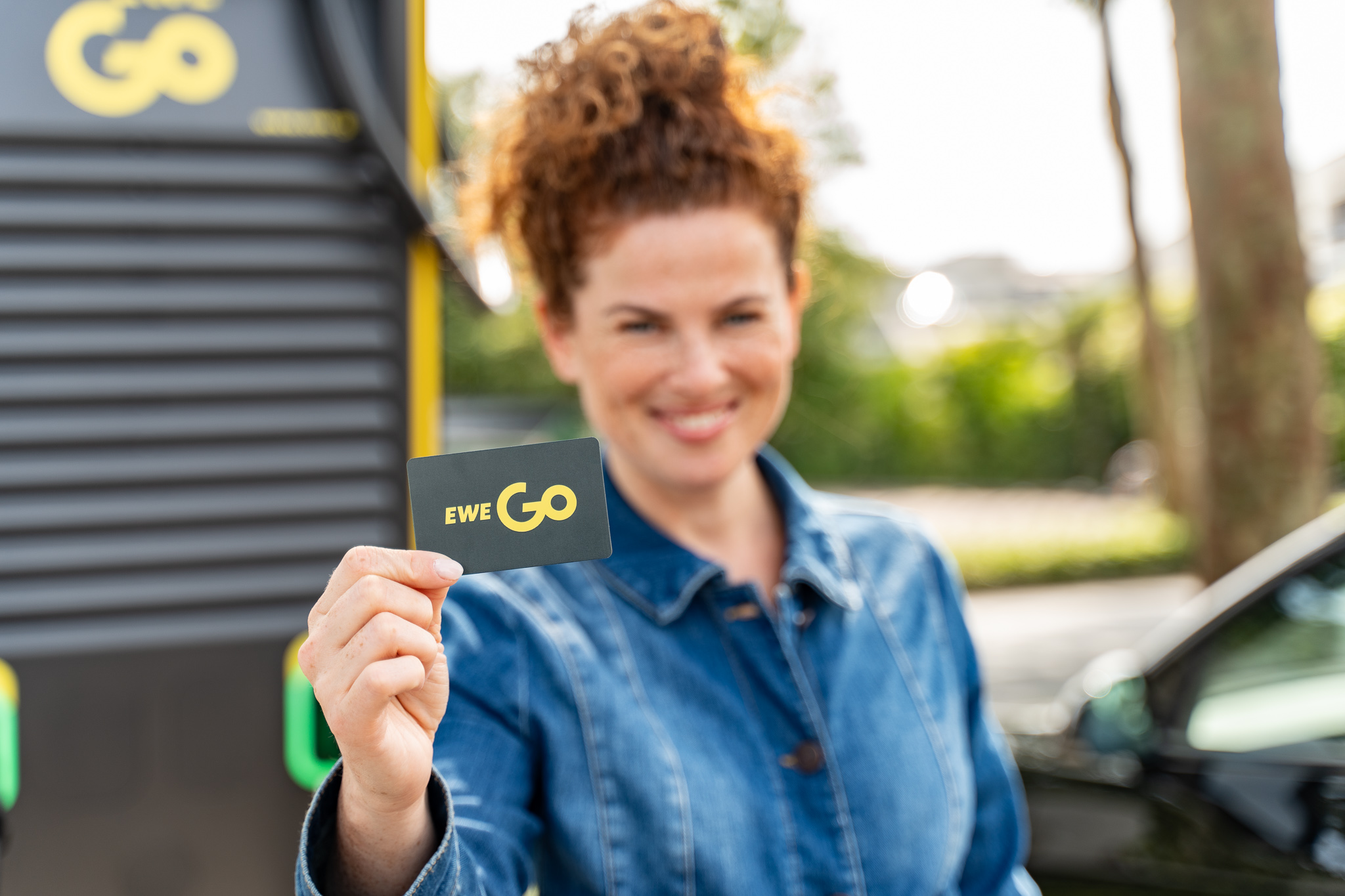 Das Bild zeigt eine Frau, die eine EWE Go Ladekarte in die Kamera hält. Die Person steht vor einer Ladestation für E-Autos. Der Hintergrund ist leicht verschwommen, wodurch die EWE Go Ladekarte und die Hand der Person hervorgehoben werden.