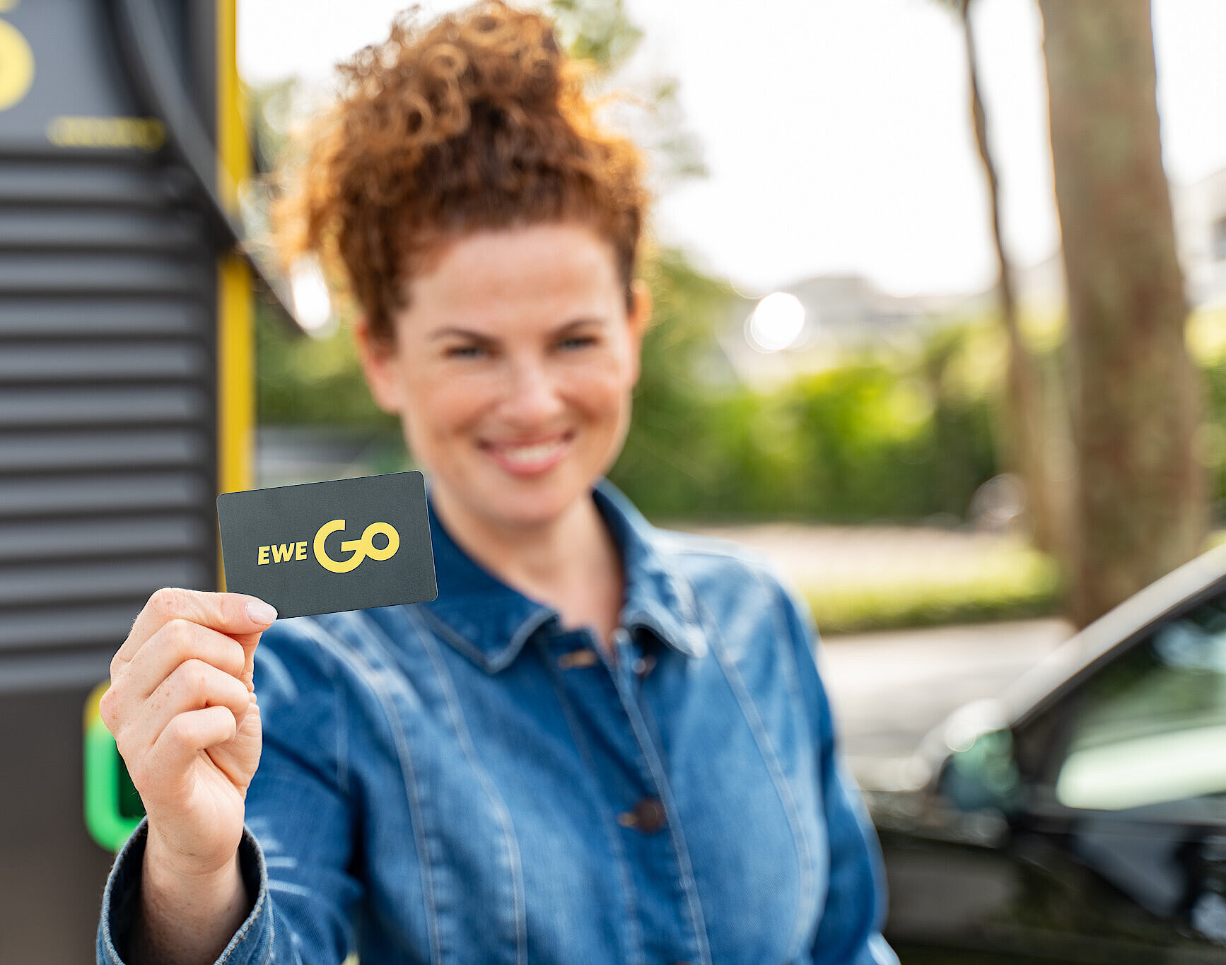 Das Bild zeigt eine Frau, die eine EWE Go Ladekarte in die Kamera hält. Die Person steht vor einer Ladestation für E-Autos. Der Hintergrund ist leicht verschwommen, wodurch die EWE Go Ladekarte und die Hand der Person hervorgehoben werden.